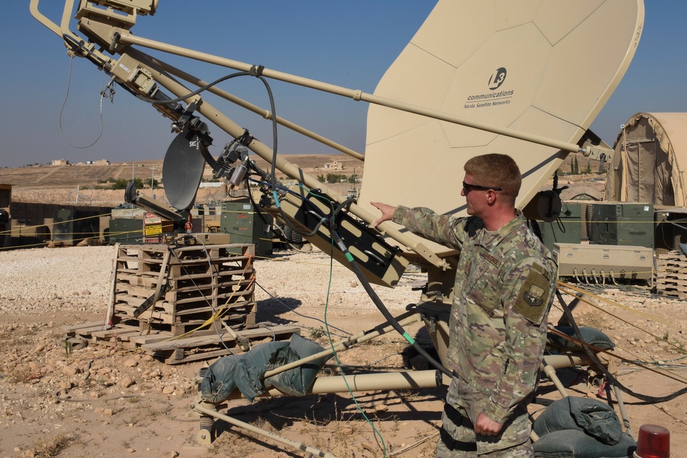 A military soldier in camouflage uniform is working with large satellite communication equipment at what appears to be a forward operating base in an arid desert environment. The primary dish is branded 'L3 Communications / Norsat Satellite Networks' and is mounted on a tan-colored support structure. A second person is partially visible on the ground near the base of the equipment.