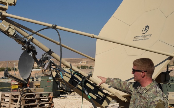 A military soldier in camouflage uniform is working with large satellite communication equipment at what appears to be a forward operating base in an arid desert environment. The primary dish is branded 'L3 Communications / Norsat Satellite Networks' and is mounted on a tan-colored support structure. A second person is partially visible on the ground near the base of the equipment.
