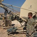 A military soldier in camouflage uniform is working with large satellite communication equipment at what appears to be a forward operating base in an arid desert environment. The primary dish is branded 'L3 Communications / Norsat Satellite Networks' and is mounted on a tan-colored support structure. A second person is partially visible on the ground near the base of the equipment.