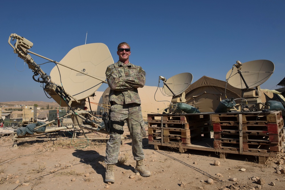 A military service member in camouflage uniform stands with arms crossed in front of multiple satellite communication dishes at what appears to be a forward operating base in an arid environment. The setting includes makeshift wooden pallet platforms supporting satellite equipment, tents, and surrounding desert terrain with visible structures in the background. The soldier appears relaxed and is smiling for the camera.