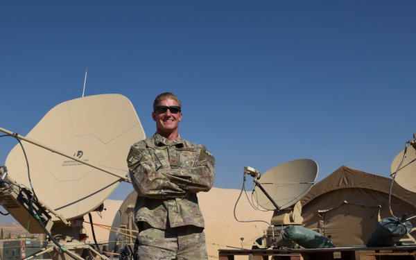 A military service member in camouflage uniform stands with arms crossed in front of multiple satellite communication dishes at what appears to be a forward operating base in an arid environment. The setting includes makeshift wooden pallet platforms supporting satellite equipment, tents, and surrounding desert terrain with visible structures in the background. The soldier appears relaxed and is smiling for the camera.