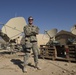 A military service member in camouflage uniform stands with arms crossed in front of multiple satellite communication dishes at what appears to be a forward operating base in an arid environment. The setting includes makeshift wooden pallet platforms supporting satellite equipment, tents, and surrounding desert terrain with visible structures in the background. The soldier appears relaxed and is smiling for the camera.