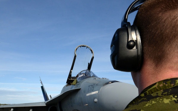 A military ground crew member wearing digital camouflage uniform and hearing protection headset observes a fighter jet taxiing on a runway. The aircraft, marked with the number '778', appears to be an F/A-18 Hornet with its canopy open. The setting is a military airfield with clear skies and tree line visible in the background.