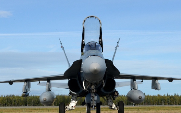 A military jet fighter, consistent with an F/A-18 Hornet configuration, is taxiing on a runway while a ground crew member in camouflage uniform with a yellow reflective vest signals with hand gestures. The aircraft is viewed head-on, revealing twin engines, external fuel tanks, and wing-tip missiles. The setting appears to be a northern airbase surrounded by boreal forest under clear skies.
