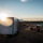 A worker in high-visibility safety gear and a hard hat is preparing or inspecting a fixed-wing UAV (drone) on a work table at an outdoor field site during golden hour. A white enclosed cargo trailer is parked nearby, likely serving as a mobile operations base. The scene appears to be a drone survey or mapping operation at a construction or agricultural site.