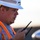 A male worker wearing a white hard hat with 'BNSF' branding and an orange high-visibility safety vest is holding a two-way radio/walkie-talkie. He appears to be communicating or listening intently, viewed from a side profile angle. The background is blurred, suggesting an outdoor industrial or rail yard environment.