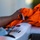 A close-up photograph showing a human hand placing or handling a crumpled orange athletic jersey or sports shirt on top of a white surface, likely a vehicle or equipment. The fabric appears to be a perforated mesh material typical of sports uniforms, with dark accent colors visible. The background is blurred, keeping focus on the hand and garment.