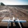 A fixed-wing UAV (unmanned aerial vehicle) with a twin-boom configuration is positioned on a dark launch pad in a flat, arid outdoor environment. The drone features white wings with orange-tipped ends and multiple propellers, suggesting a hybrid or multi-engine design. The surrounding terrain consists of bare dirt, gravel embankments, and vehicle tire tracks, with sparse trees visible on the horizon.