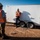 Two construction or site safety workers are having a conversation near a white enclosed trailer labeled 'EX Series' at what appears to be a desert or arid construction site. Both individuals are wearing orange high-visibility safety vests and white hard hats. One worker is leaning against the trailer while holding a tablet or clipboard, while the other stands facing them.