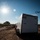 An outdoor scene in an arid, open landscape showing a white enclosed cargo trailer parked on a dirt road, with a fixed-wing drone visible in the upper right portion of the sky. A person wearing a high-visibility orange vest is partially visible beside the trailer. The image is backlit by a bright sun, creating strong lens flare and contrast.