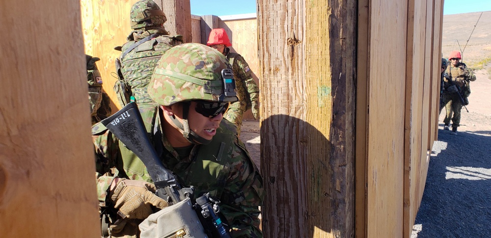Military personnel in camouflage uniforms are conducting a tactical training exercise at what appears to be a mock urban combat facility in a desert environment. Soldiers are performing a building entry or room-clearing drill through a wooden doorframe structure. The uniforms and equipment suggest Japanese Ground Self-Defense Force (JGSDF) personnel, possibly during a joint training exercise.