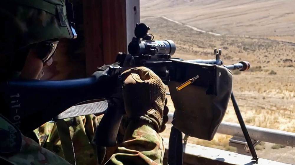 A soldier in camouflage uniform and helmet is positioned at a firing point, aiming a light machine gun equipped with an optical scope and bipod. The weapon has a visible ammunition feed tray and the soldier is wearing tactical gloves. The background shows an arid, open terrain consistent with a training range or operational environment.
