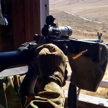 A soldier in camouflage uniform and helmet is positioned at a firing point, aiming a light machine gun equipped with an optical scope and bipod. The weapon has a visible ammunition feed tray and the soldier is wearing tactical gloves. The background shows an arid, open terrain consistent with a training range or operational environment.