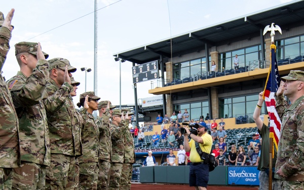 A military re-enlistment or oath ceremony is taking place on the field of a minor league baseball stadium. A group of soldiers in Army Combat Uniforms (ACU/OCP camouflage) are raising their right hands in unison before an officer standing next to a U.S. flag on a ceremonial pole. The event is being documented by a videographer and observed by stadium spectators in the background.