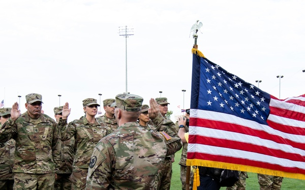 A military oath ceremony is taking place on a baseball field, with multiple soldiers in Army Combat Uniforms (ACU/OCP camouflage pattern) raising their right hands in what appears to be a reenlistment or enlistment ceremony. A large American flag with gold fringe is prominently displayed on a ceremonial pole, held by a civilian. The ceremony is being conducted by an officer facing the group, with stadium lights and bleachers visible in the background.