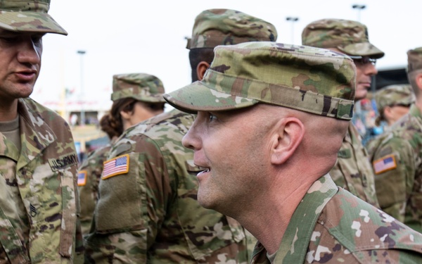 A group of U.S. Army soldiers in Operational Camouflage Pattern (OCP) uniforms are gathered together, appearing to be in conversation or during a formation event. Two soldiers are prominently visible in the foreground, with name tapes reading 'LATHAM' and partially visible 'NOM' along with 'U.S. ARMY' tape. A distinctive unit patch featuring a star and figure is visible on the right foreground soldier's left shoulder.