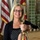 A smiling middle-aged woman with blonde hair and black-framed glasses poses indoors next to a polished brass ship's telegraph or helm fixture. She is dressed in a black cardigan over a black-and-white polka dot top and holds a cleaning cloth, suggesting she may be maintaining or displaying the artifact. The background features an American flag, what appears to be a military or organizational flag, green tiled walls, and a plaque board.