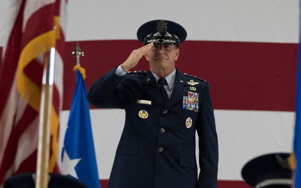 A senior U.S. Air Force general officer stands on a raised platform rendering a salute during a formal military ceremony. He is surrounded by a color guard carrying flags, with a large American flag displayed prominently in the background. The setting appears to be inside a military hangar or large indoor venue.