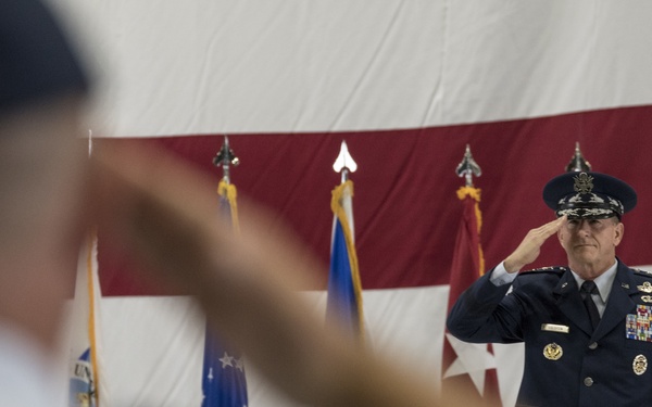 A high-ranking U.S. Air Force general officer in dress blues is rendering a salute during a formal military ceremony, likely a change of command or activation ceremony. He stands on a raised platform in front of a large American flag with military unit guidons/flags visible behind him. A blurred figure in the foreground is also saluting, suggesting a formal exchange of salutes.