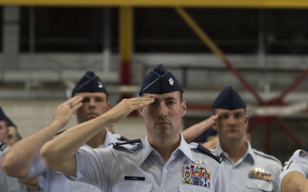 A group of United States Air Force personnel are performing a formal salute during what appears to be a military ceremony, likely a change of command or official formation. The central figure, identified by a name tag reading 'KING,' wears a Lieutenant Colonel rank insignia (silver oak leaf) and multiple service ribbons. The event is taking place inside a large hangar or indoor military facility.