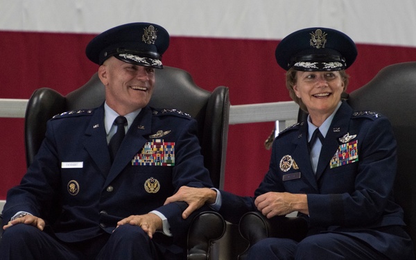 Two senior United States Air Force officers in dress blue uniforms are seated in formal leather chairs on a raised platform or stage, both smiling and appearing to share a lighthearted moment. Both individuals wear general officer rank insignia (four stars visible on shoulder boards) along with extensive medal and ribbon arrays, pilot wings, and service badges. An American flag is partially visible in the background, suggesting a formal military ceremony.