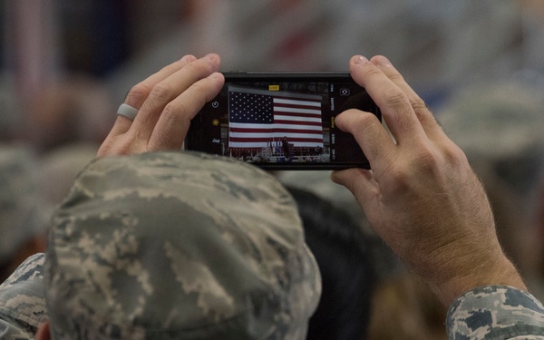 A military service member in U.S. Air Force Airman Battle Uniform (ABU) digital camouflage is photographing an American flag using a smartphone held horizontally above their head. The phone screen clearly displays the U.S. flag being captured, suggesting a formal military ceremony or event. The background shows additional uniformed personnel seated in a crowd, slightly out of focus.