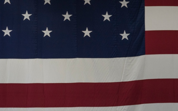 A senior U.S. Army officer in dress uniform stands at a podium delivering a speech during a formal military ceremony. He is positioned in front of a massive American flag that dominates the background, with multiple military service flags arranged behind him on staffs. The setting appears to be an indoor venue such as a hangar or large ceremonial hall.