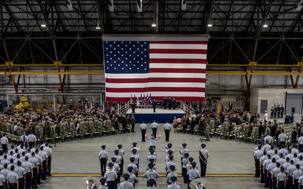 A large military ceremony is taking place inside an aircraft hangar, with uniformed personnel arranged in formal formation facing a raised stage. A large American flag is prominently displayed as a backdrop on the stage where officials are gathered. The scene appears to be a significant military event such as a change of command, activation ceremony, or similar formal military occasion.
