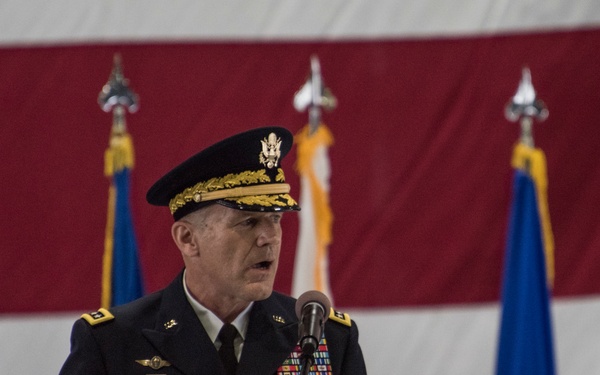 A U.S. Army general officer named Lyons, wearing dress blues with a general officer's cap, speaks at a podium during a formal military ceremony. He is positioned in front of a large American flag backdrop with multiple military service flags visible behind him. His uniform displays the 82nd Airborne Division patch, aviator wings, and numerous service ribbons.
