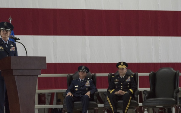 A formal military ceremony is taking place inside what appears to be a large hangar or indoor venue. A senior officer in US Air Force dress uniform is delivering a speech at a podium, while three other high-ranking officers — two in Air Force blue and one in Army green — are seated on a raised stage behind them. A large American flag serves as a backdrop, and a uniformed audience is seated in the foreground.