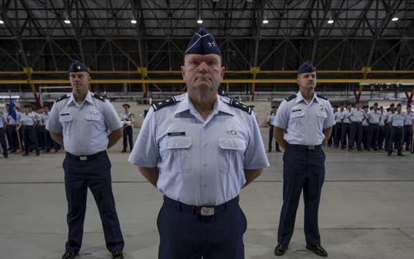 A formal military ceremony is taking place inside a large aircraft hangar, featuring personnel in United States Air Force dress uniforms. Three senior officers stand prominently in the foreground, with the central figure appearing to hold a higher rank based on insignia visible on his shoulders. Rows of uniformed airmen are assembled in formation in the background, with at least one military aircraft partially visible.