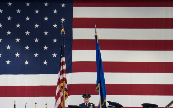 A formal U.S. Air Force color guard ceremony is taking place in front of a large American flag backdrop, likely inside a hangar or large indoor venue. Personnel in dress blue uniforms are carrying the U.S. national flag, an Air Force flag, and ceremonial rifles. A senior officer stands elevated on a stage in the background, observing the proceedings near a podium.