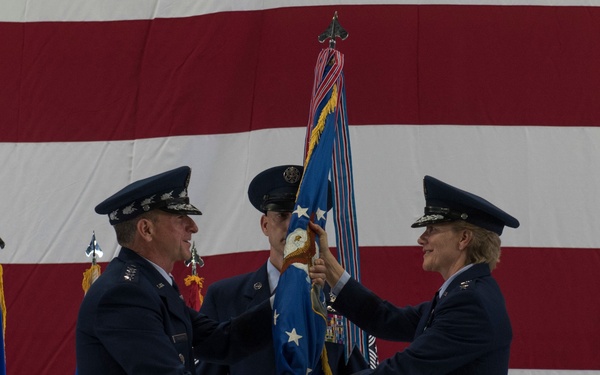 A military change of command ceremony is taking place, with two high-ranking U.S. Air Force officers exchanging a unit guidon (flag) on a formal stage. A large American flag serves as the backdrop, and a third uniformed officer stands at attention in the background. The ceremony follows traditional military protocol for transferring command authority.