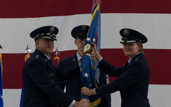 A formal U.S. Air Force change of command ceremony is taking place in front of a large American flag backdrop. Two senior officers, including one female general, are exchanging a unit guidon (flag), which is the traditional symbol of command transfer. The ceremony appears to be held in a military hangar with additional flags and personnel present.