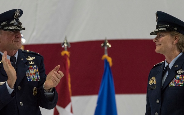 Two high-ranking United States Air Force officers are participating in what appears to be a formal military ceremony, likely a change of command or promotion event. The male officer on the left is applauding the female officer on the right, both wearing dress blue uniforms with extensive medal and ribbon arrays. The backdrop includes a large American flag and military unit flags.
