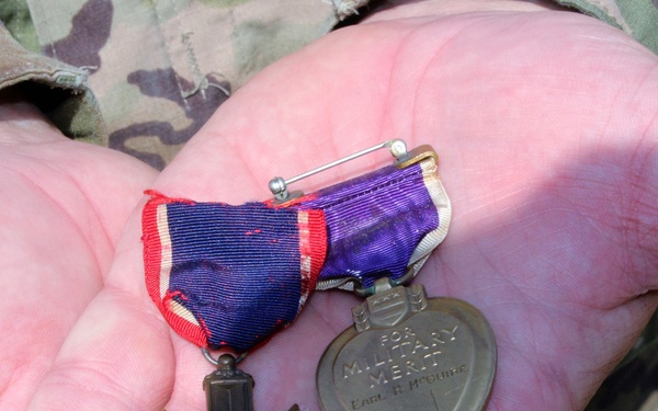 A person wearing US Army camouflage (OCP/MultiCam) uniform is holding two military medals in their open palm. The medals include a Distinguished Service Cross (or similar cross-shaped medal) with a red, white, and blue ribbon, and a Purple Heart medal with a purple and white ribbon inscribed 'FOR MILITARY MERIT' with the name 'EARL F. McGUIRE' engraved on it. Both medals appear aged and worn, suggesting they are historical artifacts.