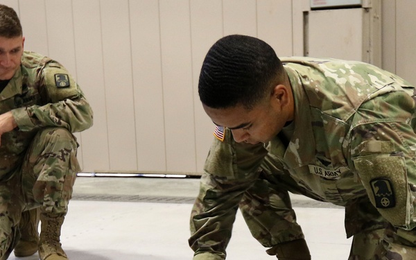 Two U.S. Army soldiers in Operational Camouflage Pattern (OCP) uniforms are kneeling on a light-colored floor inside what appears to be a military facility or hangar. The soldier in the foreground is actively handling a black cable or hose, while the second soldier observes from behind. Both wear American flag patches and unit insignia on their uniforms.