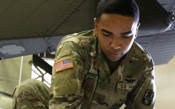 A U.S. Army soldier named Perez, wearing Operational Camouflage Pattern (OCP) uniform, is crouched down on a hangar floor inspecting or handling what appears to be a rotor blade or aircraft component. The setting is an aviation maintenance hangar, with a military aircraft visible overhead and yellow ground support equipment in the background. The soldier appears to be conducting a pre-flight or maintenance inspection.