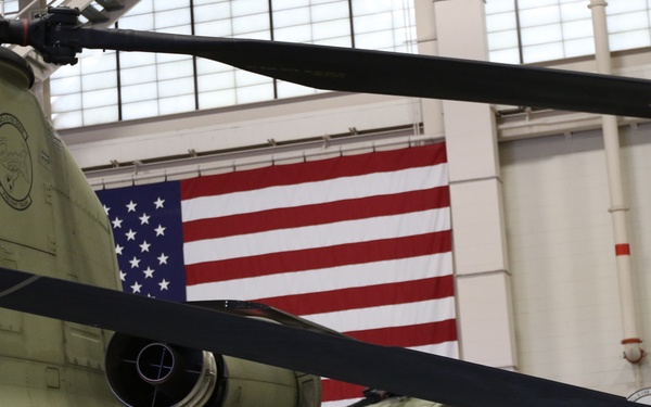 A CH-47 Chinook tandem-rotor military helicopter is photographed inside a maintenance hangar. The aircraft bears the tail number 11-08413 and is painted in olive drab/army green. An American flag and what appears to be the Virginia state flag are displayed prominently on the hangar wall in the background.