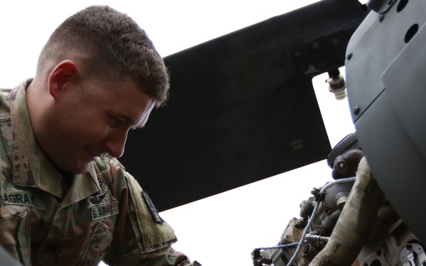 A U.S. Army soldier is performing maintenance or inspection work on what appears to be a military helicopter engine or mechanical system. The soldier is wearing Operational Camouflage Pattern (OCP) uniform with a U.S. flag patch and a name tape partially reading 'AGRA' or similar. The photo is taken from a low angle through a circular opening, likely a rotor hub or engine cowling access point.