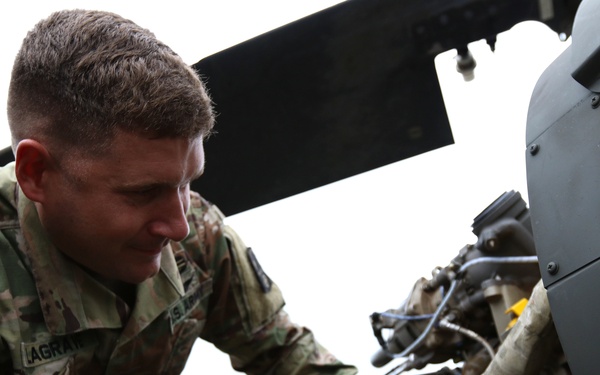 A U.S. Army soldier assigned to the 10th Mountain Division is performing maintenance or inspection work on what appears to be a military helicopter engine or mechanical system. The soldier is wearing OCP (Operational Camouflage Pattern) uniform with a U.S. flag patch and 10th Mountain Division patch visible on the left sleeve. The image is taken from a close, low angle, emphasizing the technical work being performed.