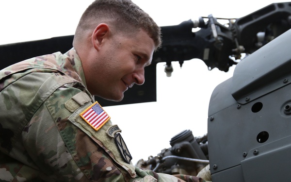 A U.S. Army soldier in OCP (Operational Camouflage Pattern) uniform is performing maintenance or inspection work on what appears to be a military helicopter. The soldier is leaning into the aircraft's mechanical components, smiling while working. An American flag patch and a unit patch are visible on the left shoulder of the uniform.