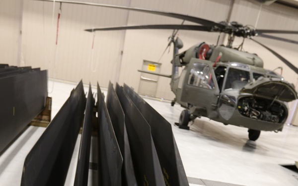 The image shows multiple helicopter rotor blades stored on a wooden rack in a maintenance hangar. The blades are dark/black in color with visible identification markings, tags, and attachment hardware at their roots. In the background, a military helicopter (appearing to be a UH-60 Black Hawk or similar variant) is visible undergoing maintenance with its cowling open.