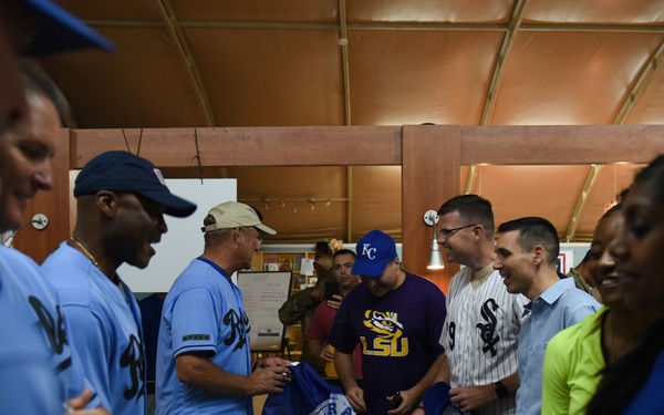A group of people are gathered around a table in what appears to be an indoor facility, handling blue jerseys or shirts. Several individuals are wearing baseball-style uniforms in light blue, while others wear casual clothing including an LSU shirt and a Chicago White Sox jersey. The scene suggests a jersey or uniform distribution event.