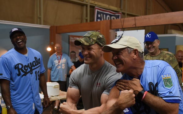 A group of people are gathered in what appears to be an indoor facility, with several individuals wearing Kansas City Royals baseball jerseys. Two men in the foreground are playfully arm-wrestling or mock-fighting while laughing, one wearing a camouflage cap and gray compression shirt, the other in a Royals jersey and tan cap. The atmosphere is festive and jovial, with onlookers smiling and laughing in the background.
