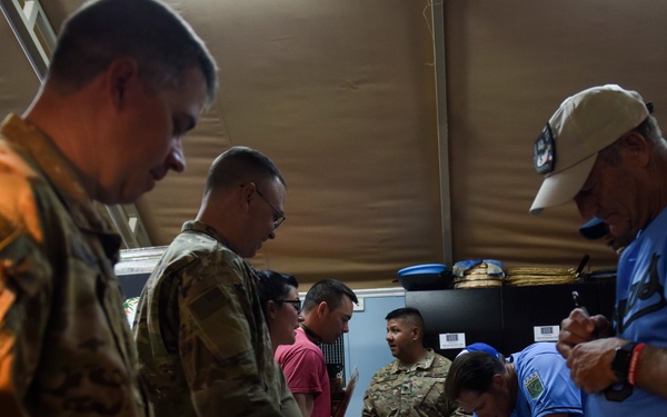 A group of military personnel in camouflage uniforms and civilians are gathered around a table in what appears to be a tent or temporary facility. They appear to be handling and signing items, possibly jerseys or shirts, on a table covered with a light-colored cloth. The setting suggests an autograph session or distribution event at a military installation.