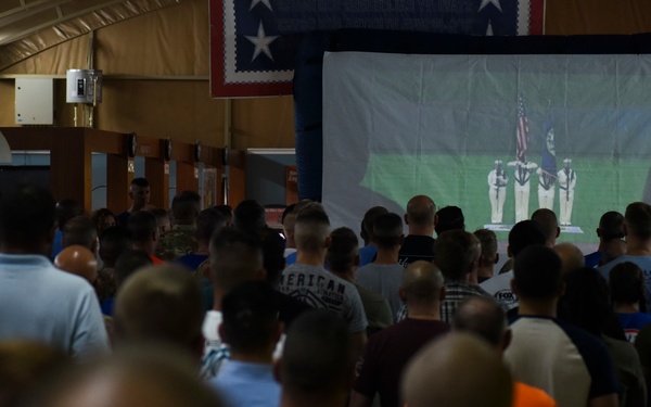 A large crowd of people, including military personnel in camouflage uniforms and civilians in casual clothing, are gathered in what appears to be a military recreation or dining facility. They are all standing and facing a large inflatable projection screen displaying what appears to be a sporting event or ceremony. A star-spangled banner or similar patriotic flag is visible hanging from the ceiling rafters.