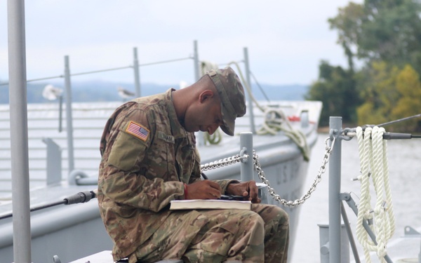 A U.S. Army soldier in MultiCam/OCP camouflage uniform is seated on the deck of a military watercraft, writing or reviewing documents on a notepad or binder. The soldier is positioned near mooring equipment including chains, ropes, and metal railings, with a body of water and tree-lined shore visible in the background. The setting appears to be a dock or pier area during daylight hours.