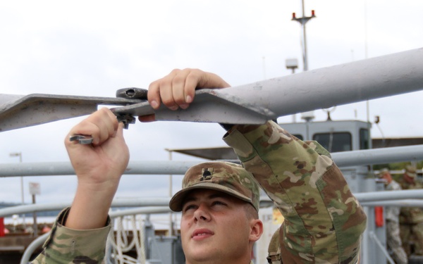 A U.S. Army soldier named LAFIN is performing maintenance or assembly work on a railing or structural component aboard a military watercraft. The soldier is wearing OCP (Operational Camouflage Pattern) uniform and using a wrench or similar hand tool. The setting appears to be a military dock or port facility with additional vessels and personnel visible in the background.