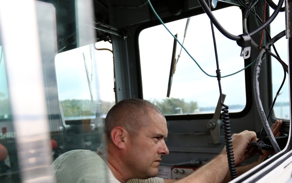 A middle-aged man with a shaved head is working inside the helm/cockpit area of a boat, appearing to perform maintenance or installation work on the controls or wiring. He is wearing a light green t-shirt and is focused on the instrument panel area. The image is taken from outside the vessel through the windshield frame, with water and trees visible in the background.